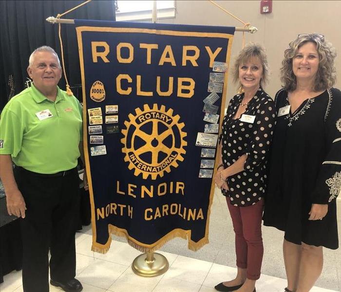 Three SERVPRO employees standing next to dark blue Lenoir Rotary Club banner