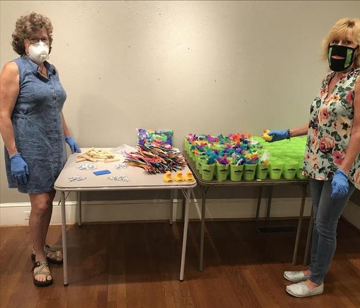 Two women wearing masks stand in front of a table with goody bags