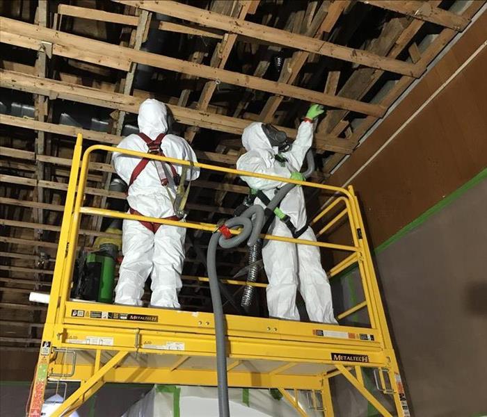 Two people in white PPE standing on a lift cleaning an exposed ceiling