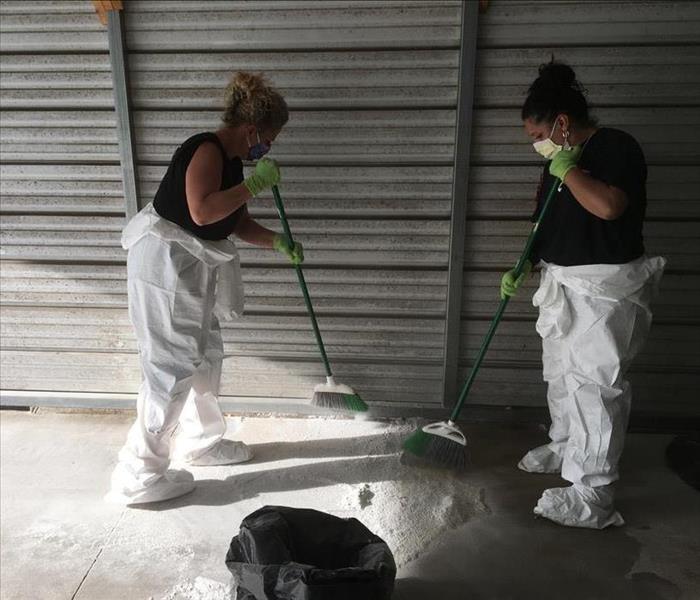 Two women wearing face masks, gloves, and partial PPE sweeping white substance on the floor